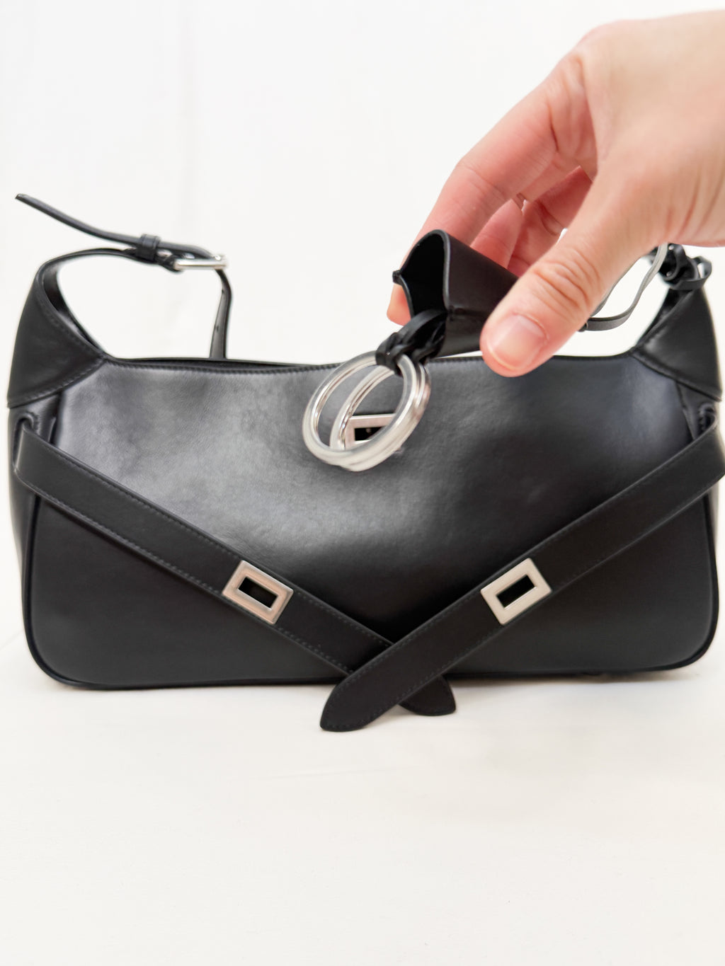 Black handbag with silver buckle and strap being adjusted by a hand on a white background